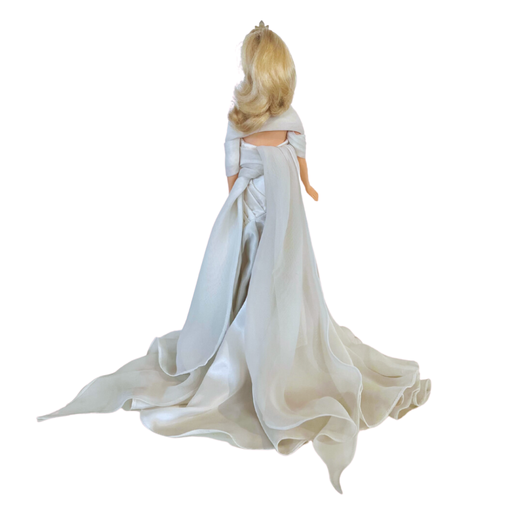 La bambola Duchess of DiamondsTM Barbie si unisce alla compagnia regale nell'acclamata Royal Jewels CollectionTM. Brilla con autentici cristalli chiari e scintillanti, squisitamente incastonati nella sua tiara, collana e orecchini. Il suo lungo abito da sera, in una tonalità perlata di charmeuse bianco, è completato da una stola di chiffon bianco sporco abbinata, che completa elegantemente l'opulento ensemble.