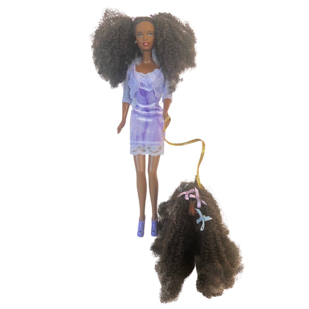 Si dice che il cane assomigli al proprio padrone ed è sicuramente il caso di questa Barbie che ha la chioma identica al pelo del suo animaletto!  Christie Barbie brilla con un abito sottoveste bordato in pizzo viola, con un cardigan coordinato rifinito in pelliccia sintetica viola e sabot viola. Sfoggia splendidi capelli ricci stretti accentati con fiocchi viola per abbinarli al suo vestito. 