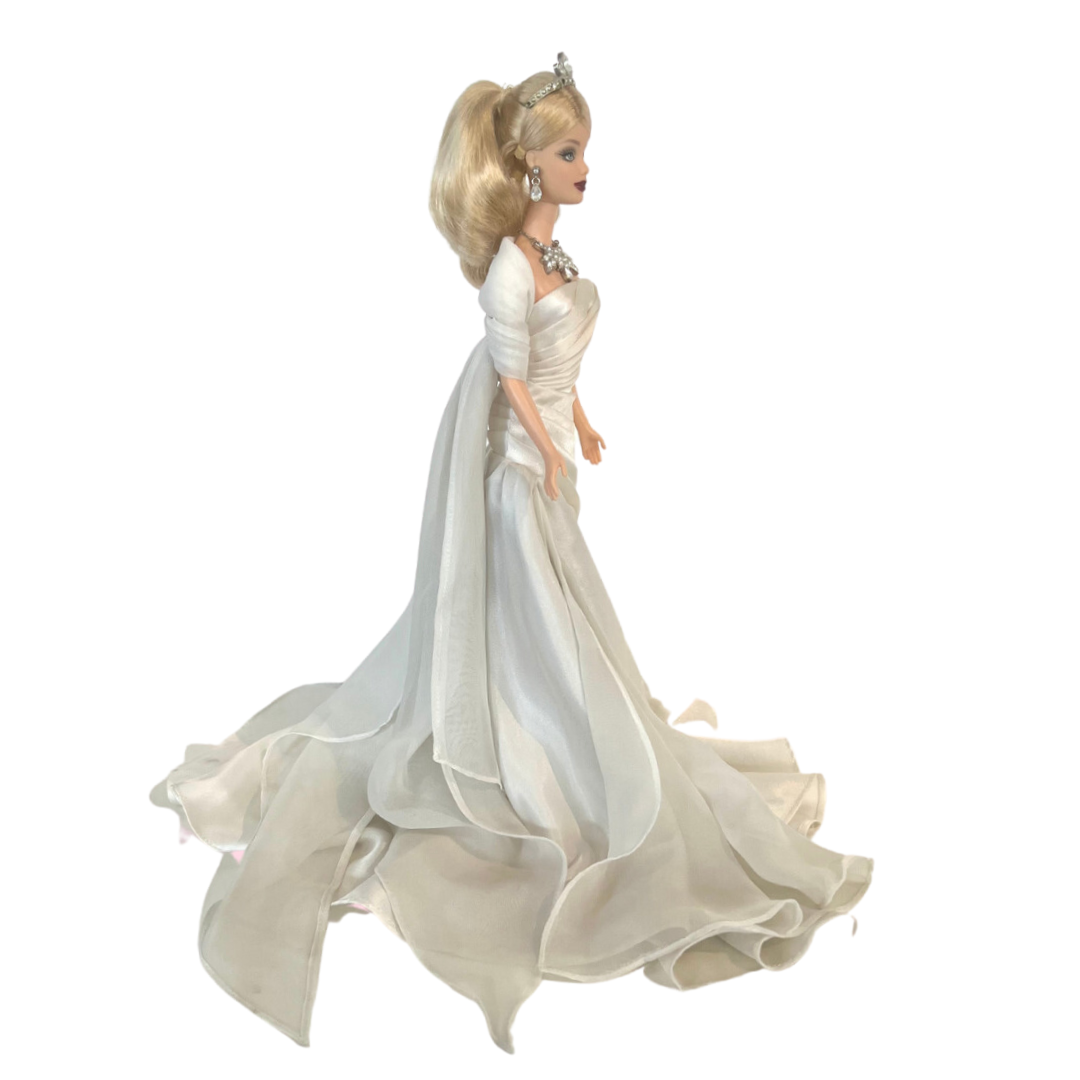 La bambola Duchess of DiamondsTM Barbie si unisce alla compagnia regale nell'acclamata Royal Jewels CollectionTM. Brilla con autentici cristalli chiari e scintillanti, squisitamente incastonati nella sua tiara, collana e orecchini. Il suo lungo abito da sera, in una tonalità perlata di charmeuse bianco, è completato da una stola di chiffon bianco sporco abbinata, che completa elegantemente l'opulento ensemble.